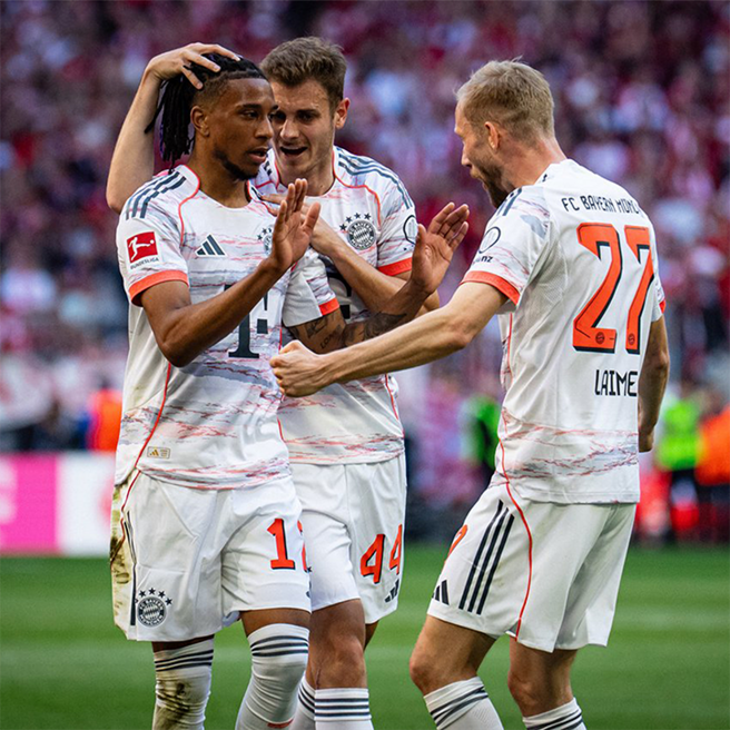 Fudbaleri Bayerna slave postignuti gol. Dva igrača su u centru pažnje, jedan se raduje i pozdravlja, dok drugi učestvuje u proslavi. U pozadini se vidi stadion sa navijačima. Atmosfera je vesela i energična.