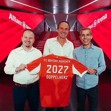 Na slici su tri muškarca koji drže crveni fudbalski dres sa natpisom "FC Bayern München" i brojem "2027". Pozadina je stadion Allianz Arena, obasjan svetlom u crvenim tonovima, što sugeriše svečanu atmosferu.