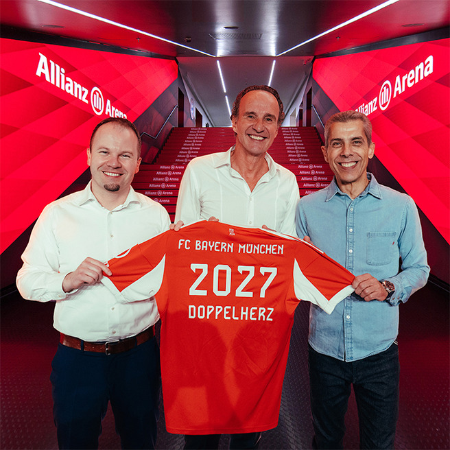 Na slici su tri muškarca koji drže crveni fudbalski dres sa natpisom "FC Bayern München" i brojem "2027". Pozadina je stadion Allianz Arena, obasjan svetlom u crvenim tonovima, što sugeriše svečanu atmosferu.